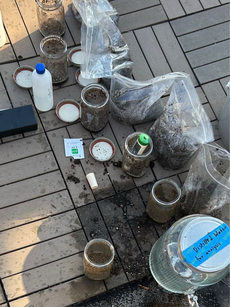 Mason jars of soil, a bottle, and a jug labeled distilled water set out on a deck